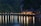 A series of charming wooden bungalows rests on a pier extending over a calm body of water. The scene is set against a backdrop of lush, forested cliffs. Warm lights illuminate the bungalows, casting long, colorful reflections onto the tranquil water's surface.