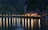 A series of charming wooden bungalows rests on a pier extending over a calm body of water. The scene is set against a backdrop of lush, forested cliffs. Warm lights illuminate the bungalows, casting long, colorful reflections onto the tranquil water's surface.