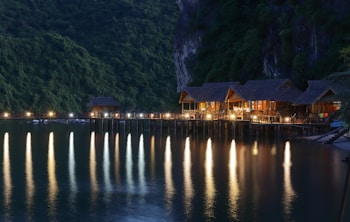 A series of charming wooden bungalows rests on a pier extending over a calm body of water. The scene is set against a backdrop of lush, forested cliffs. Warm lights illuminate the bungalows, casting long, colorful reflections onto the tranquil water's surface.