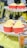 Several cupcakes with yellow frosting are arranged on a decorative white stand. The cupcakes are in red wrappers and are adorned with comic-style topper decorations featuring words like 'Boom!' and 'Bam!'. The background includes fuzzy yellow fabric and blurred decorations with a comic theme.