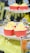 Several cupcakes with yellow frosting are arranged on a decorative white stand. The cupcakes are in red wrappers and are adorned with comic-style topper decorations featuring words like 'Boom!' and 'Bam!'. The background includes fuzzy yellow fabric and blurred decorations with a comic theme.