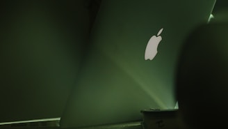 An image featuring the back of an open laptop displaying the illuminated logo. The laptop sits on a table in a dimly lit environment, with the scene predominantly bathed in shades of green.