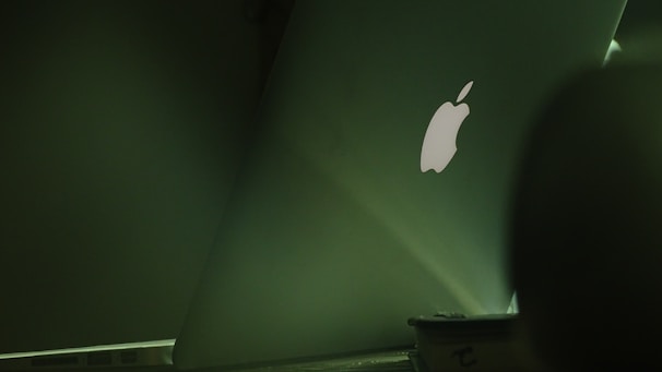 An image featuring the back of an open laptop displaying the illuminated logo. The laptop sits on a table in a dimly lit environment, with the scene predominantly bathed in shades of green.