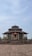 A traditional temple structure with layered roofs and a prominent central dome, built with stone and featuring multiple pillars. The entrance is adorned with steps leading up to a statue housed under a small roof. The architecture is surrounded by a paved courtyard area and sparse greenery.