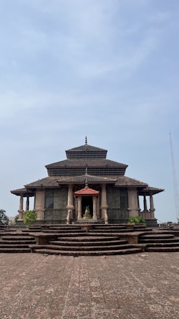 A traditional temple structure with layered roofs and a prominent central dome, built with stone and featuring multiple pillars. The entrance is adorned with steps leading up to a statue housed under a small roof. The architecture is surrounded by a paved courtyard area and sparse greenery.
