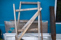 A wooden ladder leaning against a blue wall, surrounded by various wooden planks and metal rods. The wall has some peeling sections, revealing an older layer of paint. The ladder and boards appear weathered and worn.