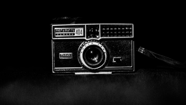 A vintage camera with a rectangular body and a prominent lens centered on a dark background. The camera has a textured surface and an attached strap on the right side.