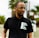 A person wearing glasses and a black polo shirt with the words 'New Breed of Deep House' printed on it, standing outdoors with palm trees blurred in the background.