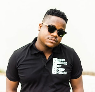 A person is standing against a light-colored background wearing a black polo shirt with the words 'NEW BREED OF DEEP HOUSE' printed on it. The person is also wearing dark sunglasses and has a short hairstyle.