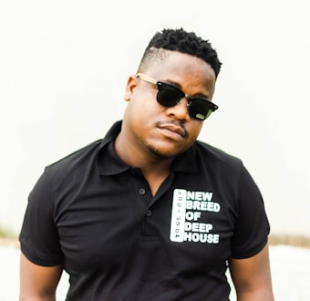 A person is standing against a light-colored background wearing a black polo shirt with the words 'NEW BREED OF DEEP HOUSE' printed on it. The person is also wearing dark sunglasses and has a short hairstyle.