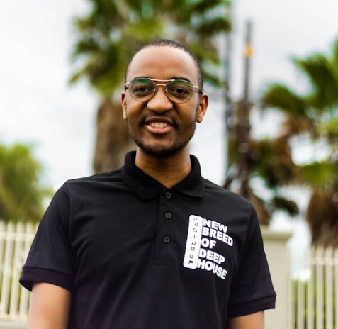 A person wearing glasses and a black polo shirt with the text 'New Breed of Deep House'. The background features blurred palm trees.