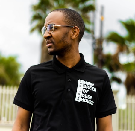 A man wearing glasses and a black polo shirt with the text 'New Breed of Deep House' printed on it stands outdoors. In the background, there are blurred palm trees and a fence, suggesting a tropical or suburban setting.