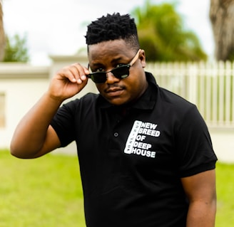 A person wearing a black polo shirt with the text 'New Breed of Deep House' printed on it, adjusting their sunglasses. The background features greenery and a fence.