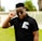 A person wearing a black polo shirt with the text 'New Breed of Deep House' printed on it, adjusting their sunglasses. The background features greenery and a fence.
