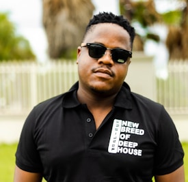 A person is wearing a black polo shirt with text saying 'New Breed of Deep House' and sunglasses. The background shows a blurred outdoor setting with trees and a fence.