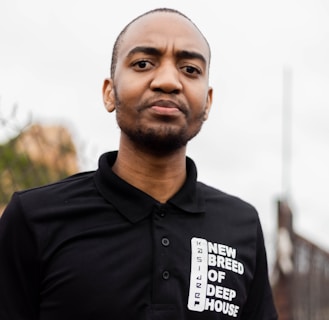 A person wearing a black polo shirt with the text 'NEW BREED OF DEEP HOUSE'. The background is blurred, suggesting an outdoor setting.