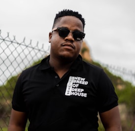 A person wearing sunglasses and a black polo shirt with white text that reads 'NEW BREED OF DEEP HOUSE'. They are standing in front of a chain-link fence with an overcast sky and some greenery in the background.