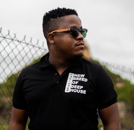 A person wearing sunglasses and a black polo shirt with the text 'New Breed of Deep House' stands in front of a chain-link fence with a slightly blurred natural background.
