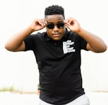 A person wearing sunglasses and a black polo shirt with white text on it stands in a confident pose. The background is plain, and the person is adjusting their sunglasses.