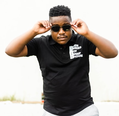A person wearing sunglasses and a black polo shirt with white text on it stands in a confident pose. The background is plain, and the person is adjusting their sunglasses.