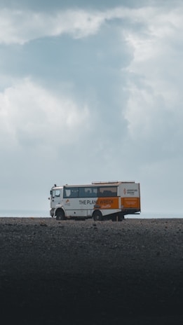 A tour bus with the text 'The Plane Wreck Shuttle' is parked on a desolate and rocky landscape. The sky is overcast with thick clouds, giving the scene a moody atmosphere.
