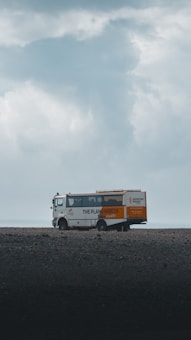 A tour bus with the text 'The Plane Wreck Shuttle' is parked on a desolate and rocky landscape. The sky is overcast with thick clouds, giving the scene a moody atmosphere.