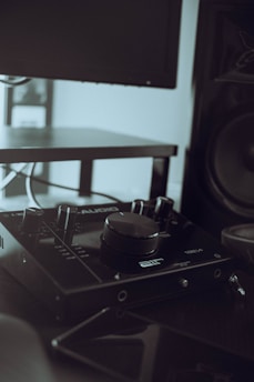 A close-up of audio equipment, specifically an audio interface with various knobs and dials, placed on a dark surface. In the background, partial views of a monitor screen and a speaker can be seen.