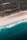 An aerial view of a coastline showing the transition from lush green land to white sandy beaches and then blue ocean waves. The landscape is marked with fields and scattered buildings visible inland.
