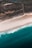 An aerial view of a coastline showing the transition from lush green land to white sandy beaches and then blue ocean waves. The landscape is marked with fields and scattered buildings visible inland.