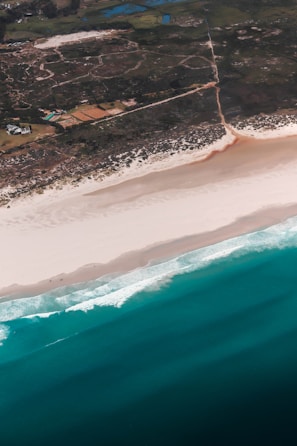 An aerial view of a coastline showing the transition from lush green land to white sandy beaches and then blue ocean waves. The landscape is marked with fields and scattered buildings visible inland.