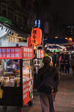 A bustling night market scene with a person walking past food stalls that are adorned with bright red signs covered in Chinese characters. A red lantern above the stall provides additional light and adds to the cultural ambiance of the setting. The background is filled with more stalls and people, suggesting a lively and busy atmosphere.
