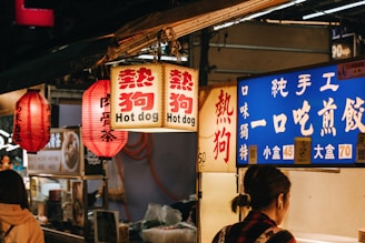 A bustling street food market with vibrant signage featuring lanterns and illuminated signs. The signs display both Chinese characters and English, mentioning hot dogs. A person, likely a customer, stands in front of the stall.