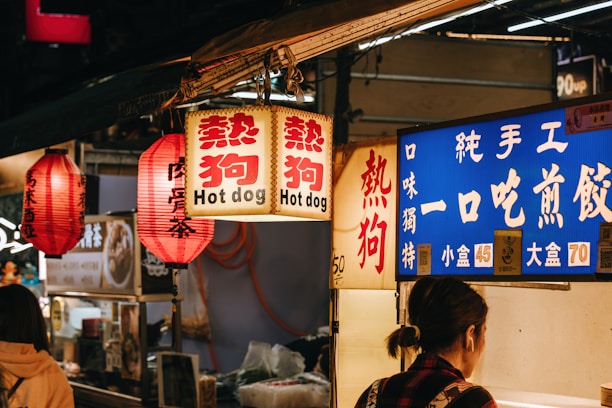 A bustling street food market with vibrant signage featuring lanterns and illuminated signs. The signs display both Chinese characters and English, mentioning hot dogs. A person, likely a customer, stands in front of the stall.