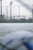 A football field with a focus on a goalpost surrounded by a chain-link fence. The ground appears frosty or covered with a thin layer of snow. The field is part of an outdoor sports facility with visible floodlights and buildings in the background. The foreground shows a blurry soccer ball, suggesting an overcast day with soft lighting.