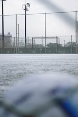 A football field with a focus on a goalpost surrounded by a chain-link fence. The ground appears frosty or covered with a thin layer of snow. The field is part of an outdoor sports facility with visible floodlights and buildings in the background. The foreground shows a blurry soccer ball, suggesting an overcast day with soft lighting.