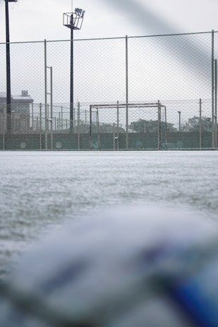 A football field with a focus on a goalpost surrounded by a chain-link fence. The ground appears frosty or covered with a thin layer of snow. The field is part of an outdoor sports facility with visible floodlights and buildings in the background. The foreground shows a blurry soccer ball, suggesting an overcast day with soft lighting.