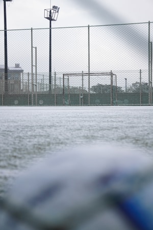 A football field with a focus on a goalpost surrounded by a chain-link fence. The ground appears frosty or covered with a thin layer of snow. The field is part of an outdoor sports facility with visible floodlights and buildings in the background. The foreground shows a blurry soccer ball, suggesting an overcast day with soft lighting.
