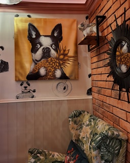 A room with a whimsical decor featuring a large painting of a black and white dog holding a pineapple against a gold background. The walls have brick and wood paneling, with illustrations of pandas in suits and a coffee cup. A plush chair with floral upholstery is in the foreground. An astronaut dog figure is placed on a shelf, and a round sunburst mirror is mounted on the wall.
