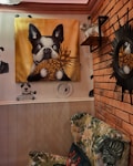 A room with a whimsical decor featuring a large painting of a black and white dog holding a pineapple against a gold background. The walls have brick and wood paneling, with illustrations of pandas in suits and a coffee cup. A plush chair with floral upholstery is in the foreground. An astronaut dog figure is placed on a shelf, and a round sunburst mirror is mounted on the wall.