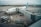 An airplane is parked at an airport gate connected by a jet bridge, with several ground vehicles and personnel nearby. The airplane is clearly marked with the airline's logo and Canadian flag. The tarmac and surrounding area appear overcast, suggesting cloudy weather.