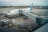 An airplane is parked at an airport gate connected by a jet bridge, with several ground vehicles and personnel nearby. The airplane is clearly marked with the airline's logo and Canadian flag. The tarmac and surrounding area appear overcast, suggesting cloudy weather.