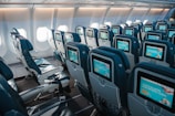An interior view of an airplane cabin showcasing rows of empty seats equipped with individual screens on the back of each seat. The cabin is well-lit by natural light coming through the airplane windows, highlighting the blue and gray color scheme of the seats.