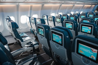 An interior view of an airplane cabin showcasing rows of empty seats equipped with individual screens on the back of each seat. The cabin is well-lit by natural light coming through the airplane windows, highlighting the blue and gray color scheme of the seats.
