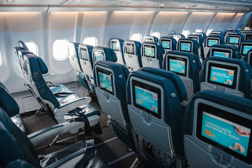 An interior view of an airplane cabin showcasing rows of empty seats equipped with individual screens on the back of each seat. The cabin is well-lit by natural light coming through the airplane windows, highlighting the blue and gray color scheme of the seats.