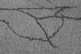 An asphalt surface with visible cracks forming irregular patterns across the pavement. The texture of the asphalt is rough, showing small pebbles and grainy details.