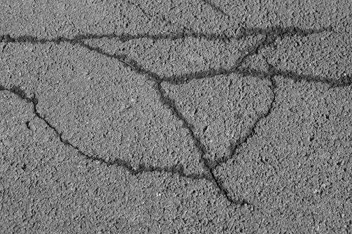 An asphalt surface with visible cracks forming irregular patterns across the pavement. The texture of the asphalt is rough, showing small pebbles and grainy details.