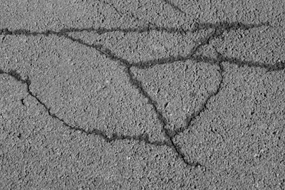 An asphalt surface with visible cracks forming irregular patterns across the pavement. The texture of the asphalt is rough, showing small pebbles and grainy details.