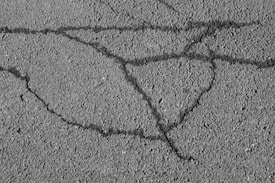 An asphalt surface with visible cracks forming irregular patterns across the pavement. The texture of the asphalt is rough, showing small pebbles and grainy details.