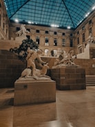 An indoor gallery with classical sculptures displayed on stone pedestals. The ceiling is made of glass panels, allowing natural light to illuminate the stone walls and floors. Statues include a human figure interacting with an animal, and other figures in dynamic poses.