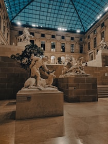 An indoor gallery with classical sculptures displayed on stone pedestals. The ceiling is made of glass panels, allowing natural light to illuminate the stone walls and floors. Statues include a human figure interacting with an animal, and other figures in dynamic poses.